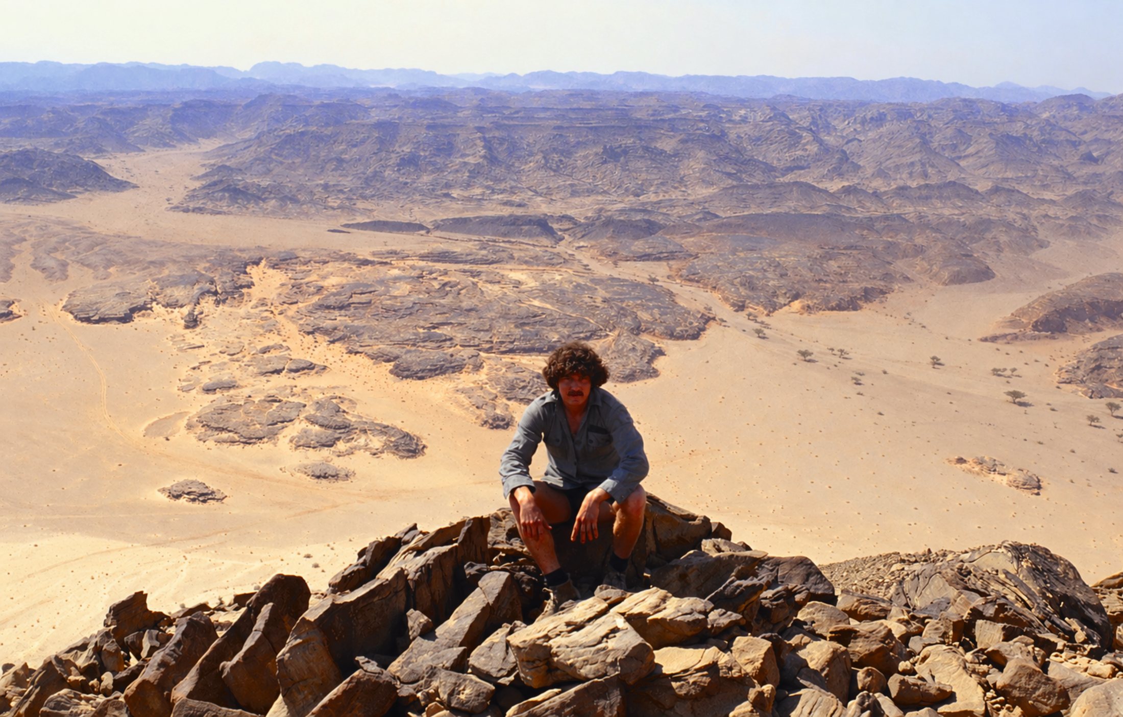 Michael crouching on a rocky ridge high above a vast desert valley and distant mountains at Jebel Milehis, Sinai, 1981